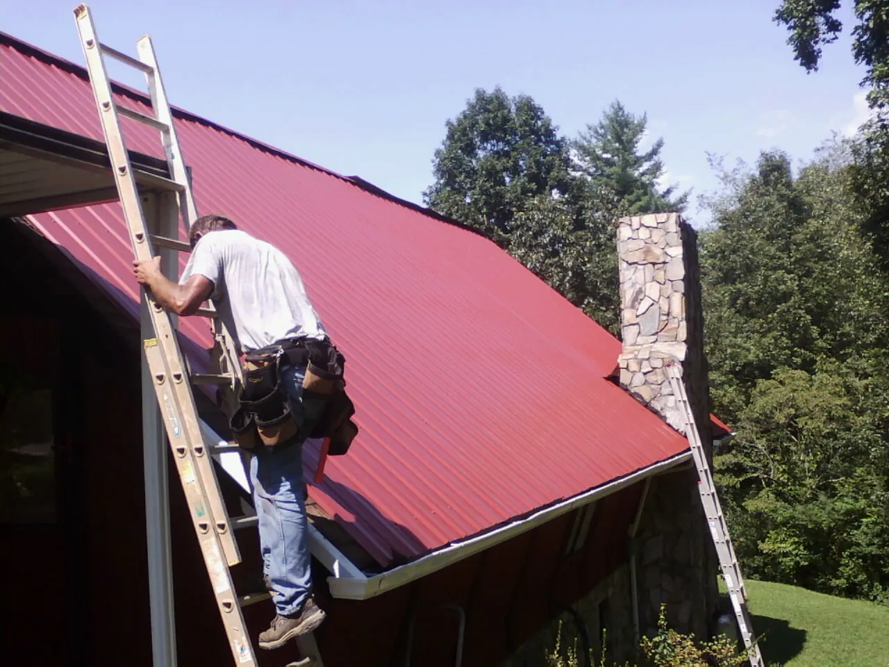 Roofer on a ladder beside a red metal roof and stone chimney — Soffit & Fascia Repair by Roofing Brothers in Cherry Valley